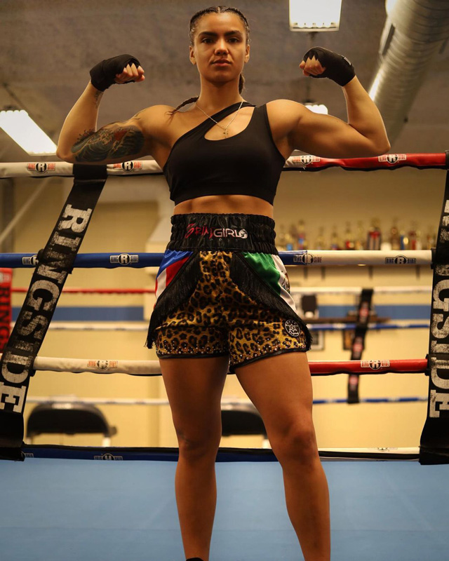 The coach stands in a boxing ring wearing boxing equipment with thier opponent in the background.